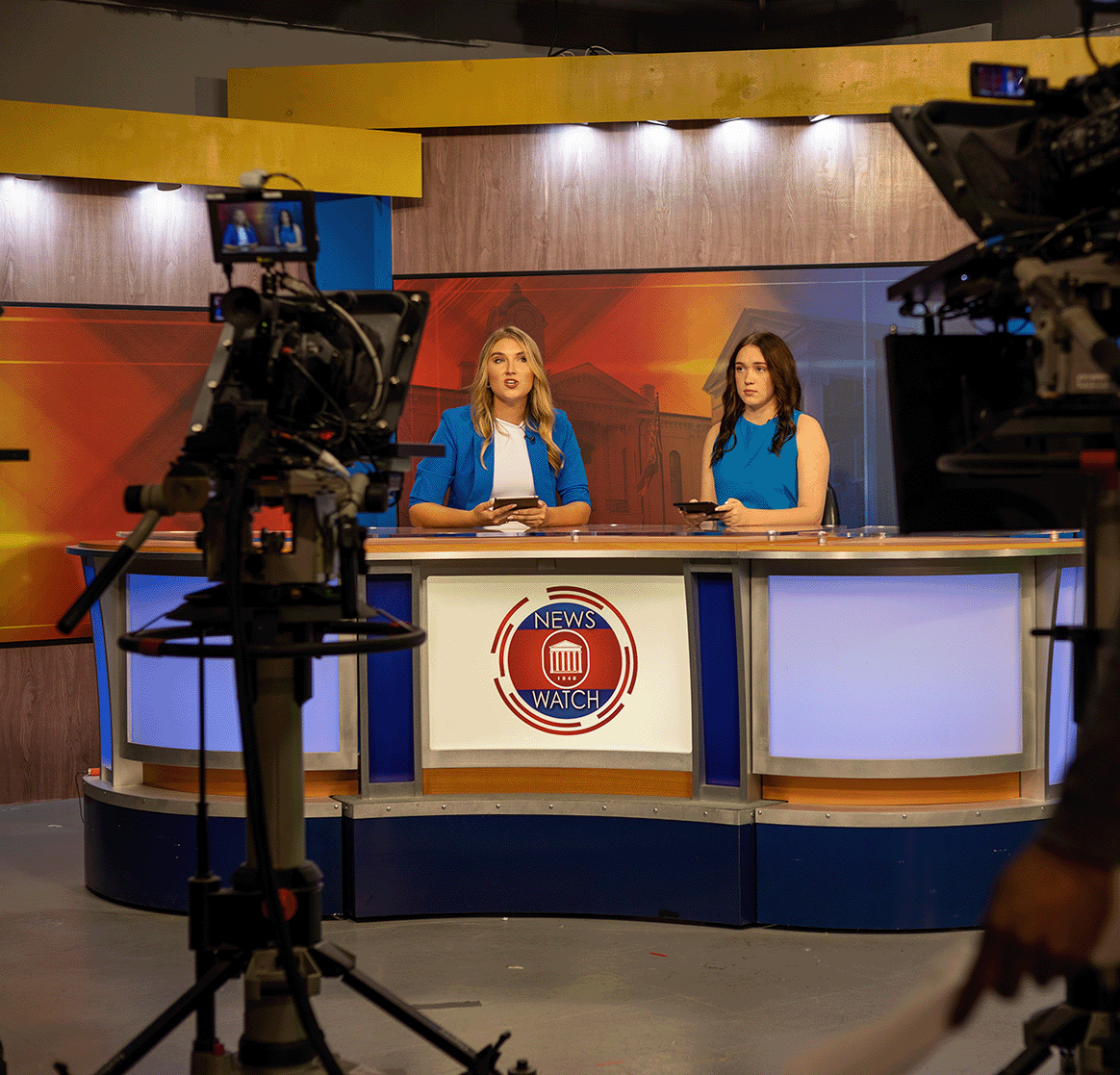 Two news anchors sit behind a news desk, talking to a camera.