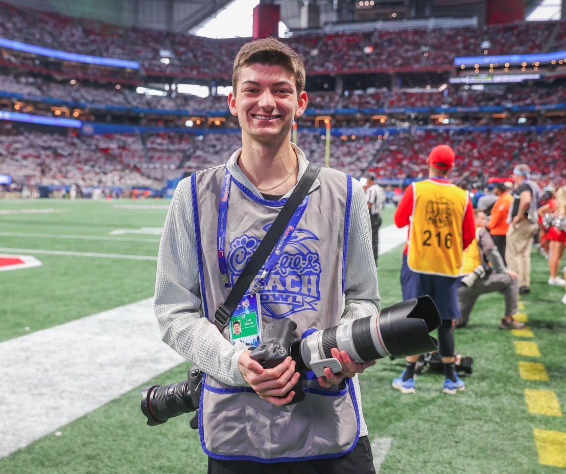 A student stands on the football sidelines while holding a camera.