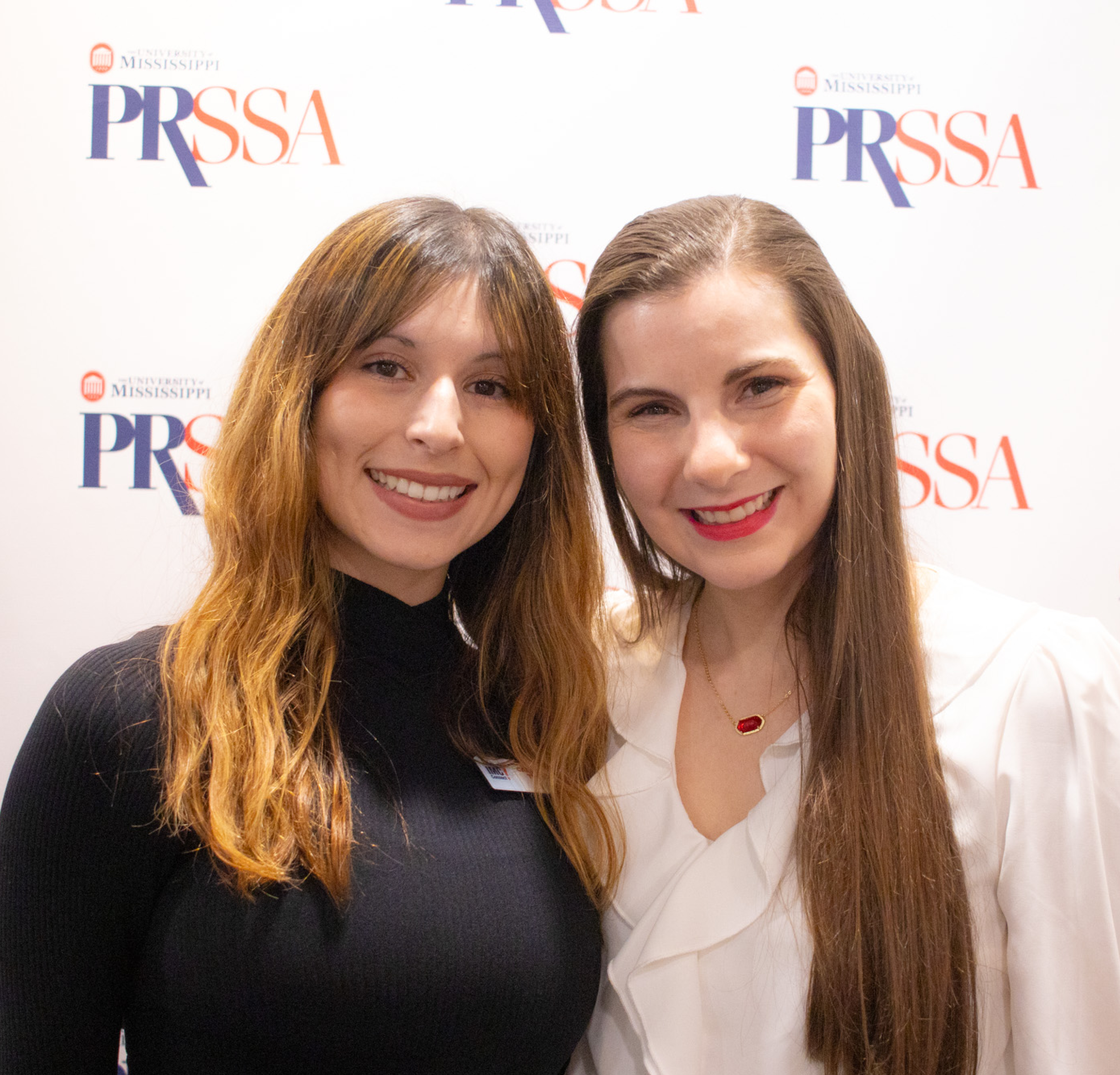 Two students pose for a photo in front of a backdrop with the PRSSA logo.