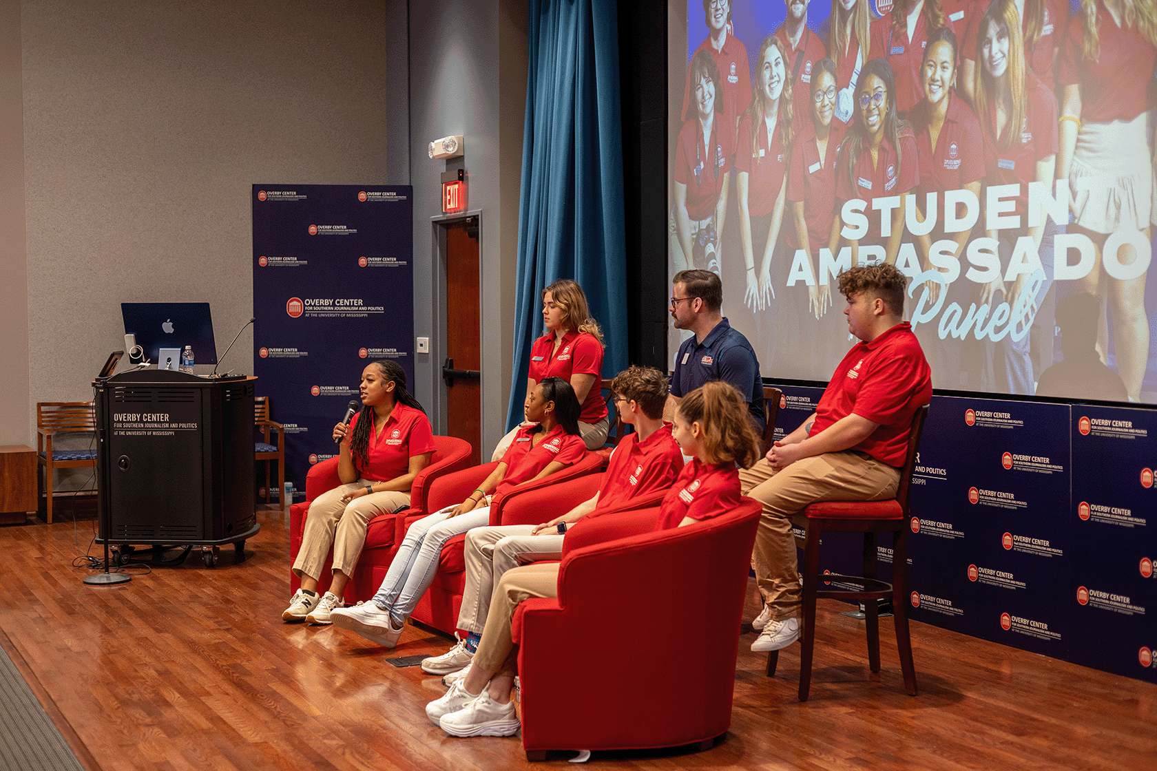 A group of students sit in front of an auditorium talking to guests.