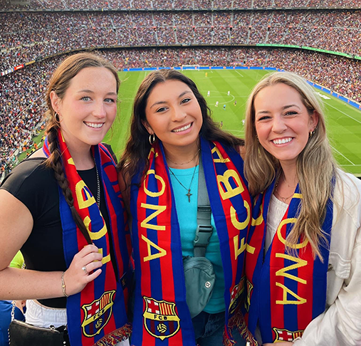 Three students pose for a photo in front of a large soccer arena.