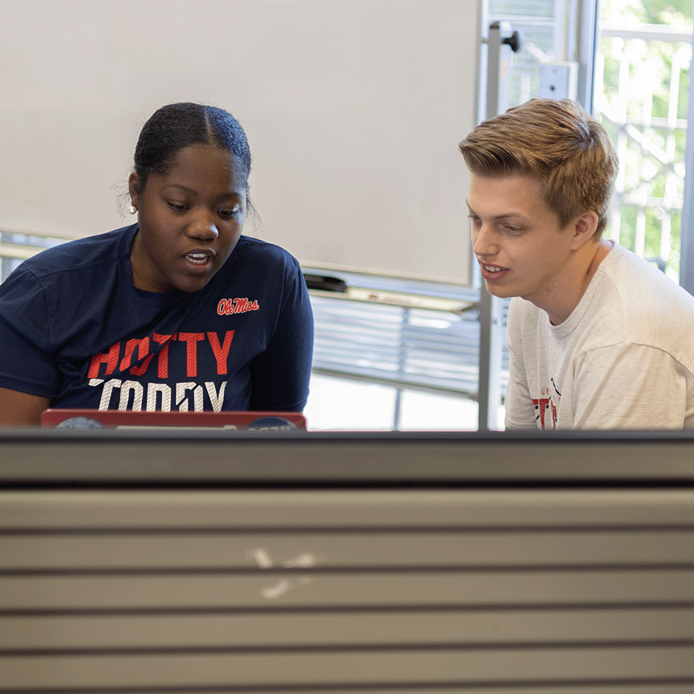 Two students talk while looking down at a laptop.