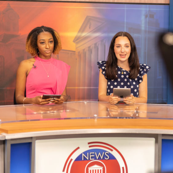 Two students sit on a news set reporting the news